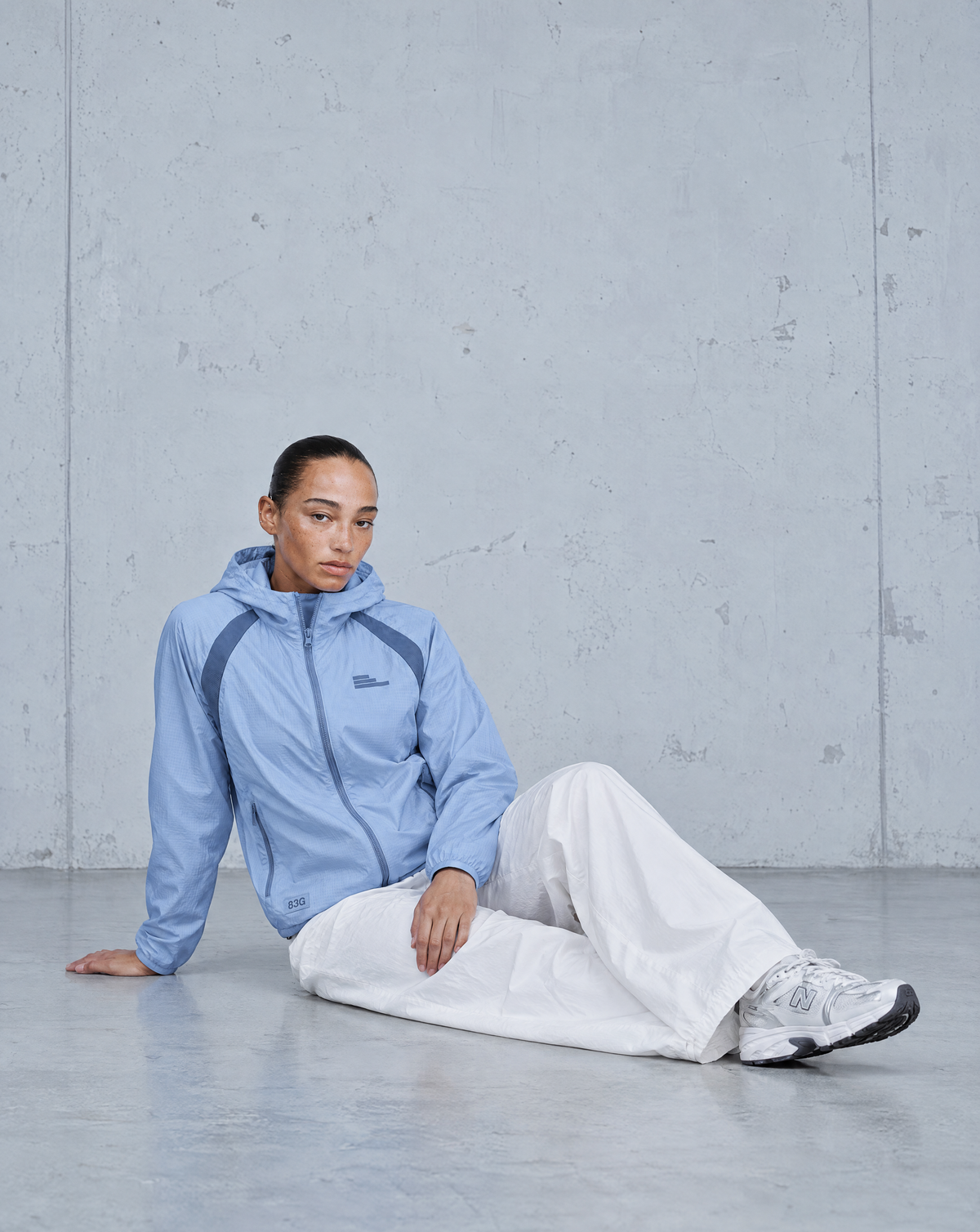 Model wearing a light blue Five O’ Five jacket and white trousers, seated against a concrete wall.